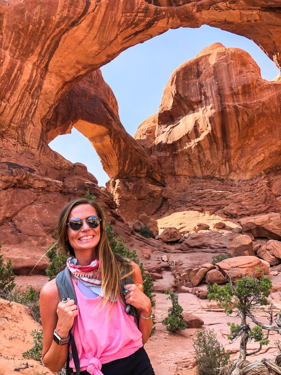 Emily, wearing a pink shirt, sunglasses, and a bandana around her neck, smiles in front of giant arches at Arches National Park.