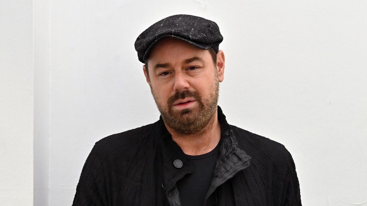 Danny Dyer stood in front of a white wall. He is wearing a black top, jacket and hat.