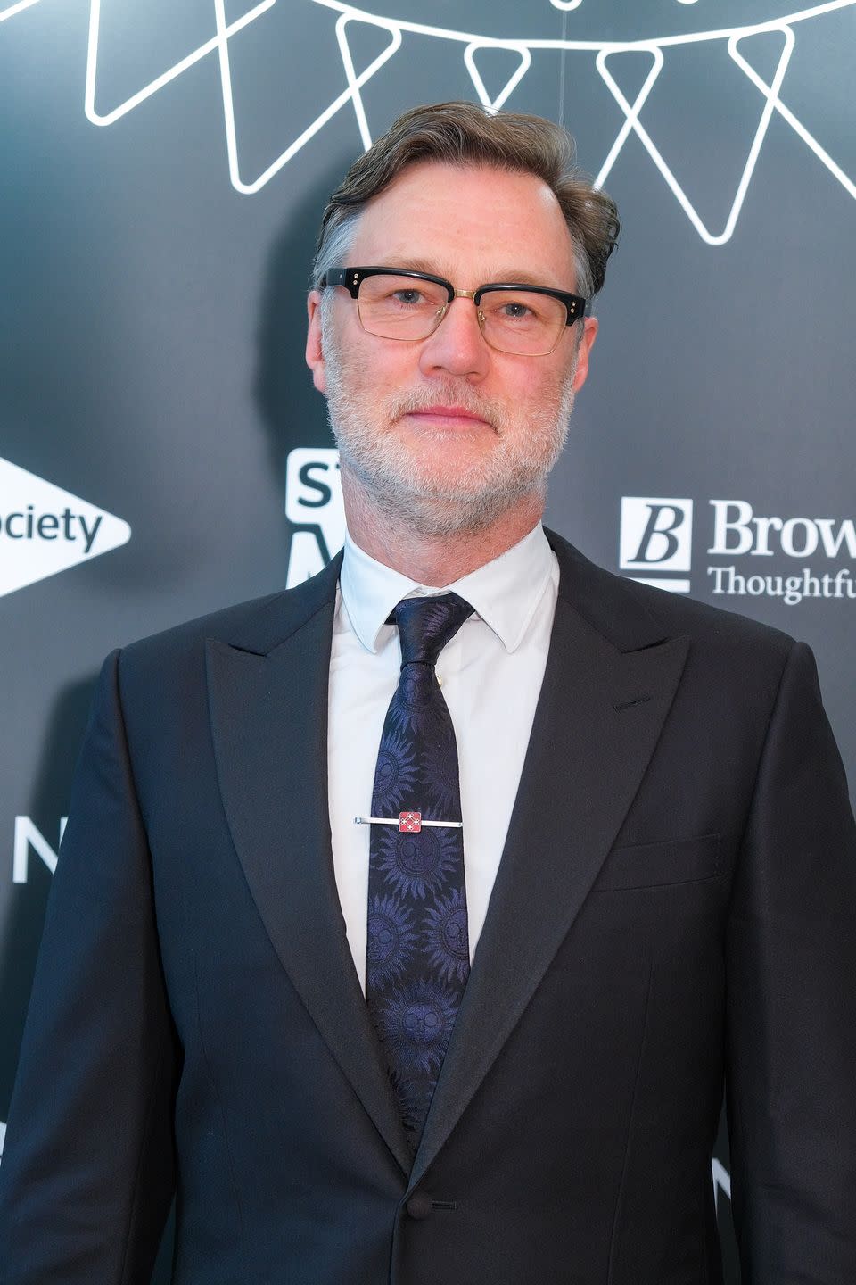 david morrissey, an older man stands looking at the camera with a neutral facial expression, he wears glasses with greying hair slicked back, a black suit jacket, white shirt and blue tie