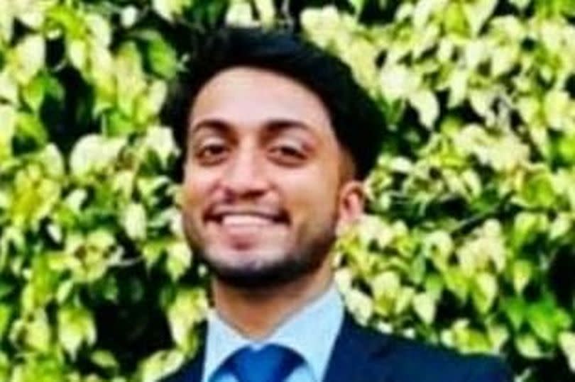 Jubal Reji Kurian in blue suit with tie, smiling, against backgrop of light green leaves