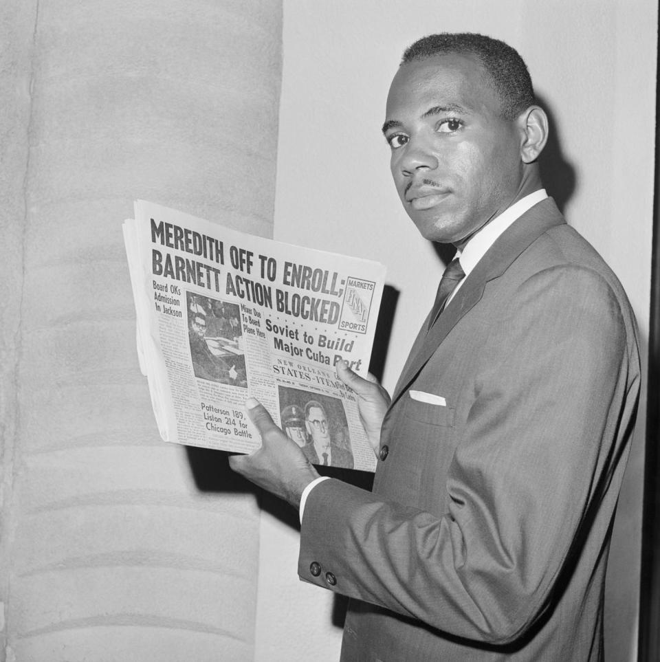 james meredith holds a newspaper while standing and looking over his left shoulder at the camera, he wears a suit jacket and tie and has a small mustache