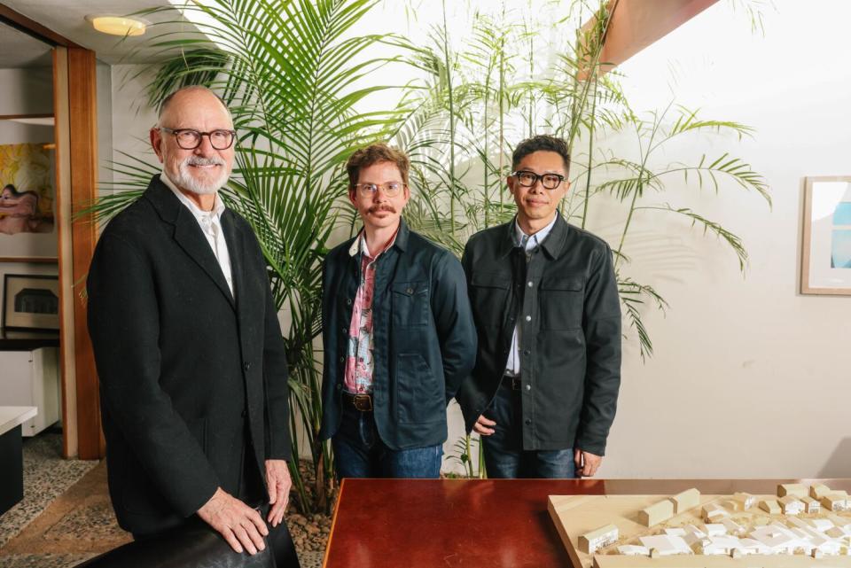 Three men stand in a conference room with a bright skylight that illuminates palm trees.