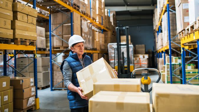 Senior male warehouse worker loading or unloading boxes from a pallet truck.