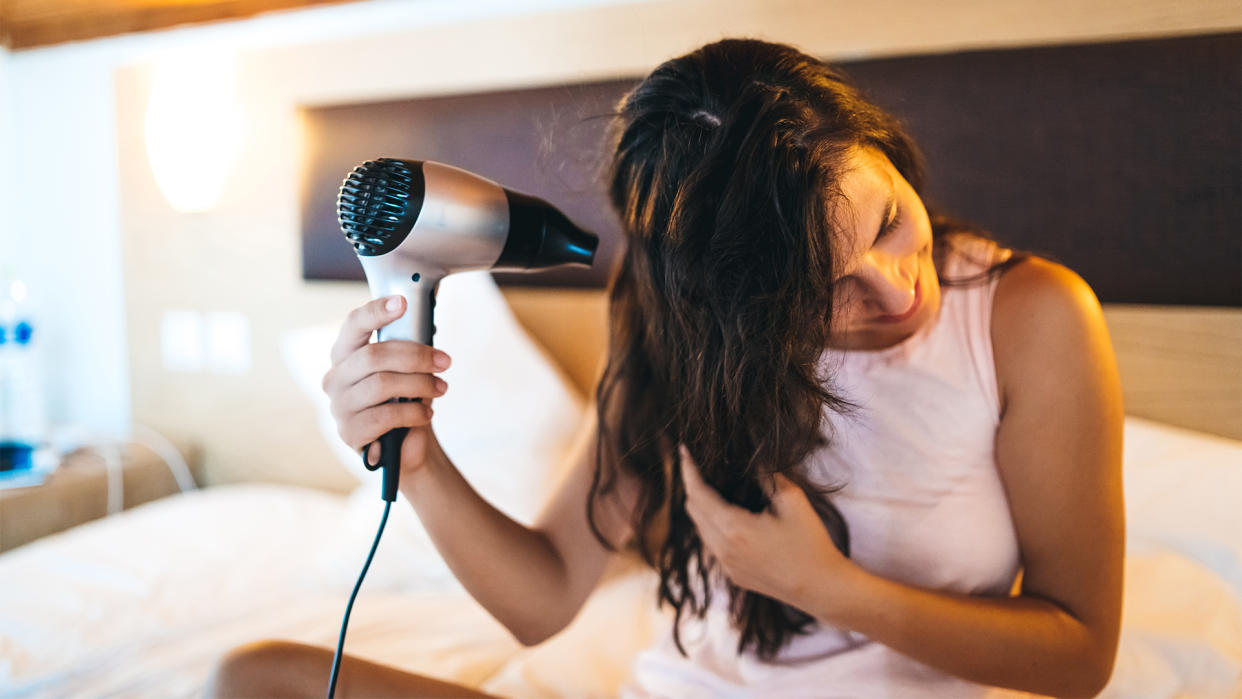  A woman who has just gotten out of the shower sits on her mattress and dries her hair. 