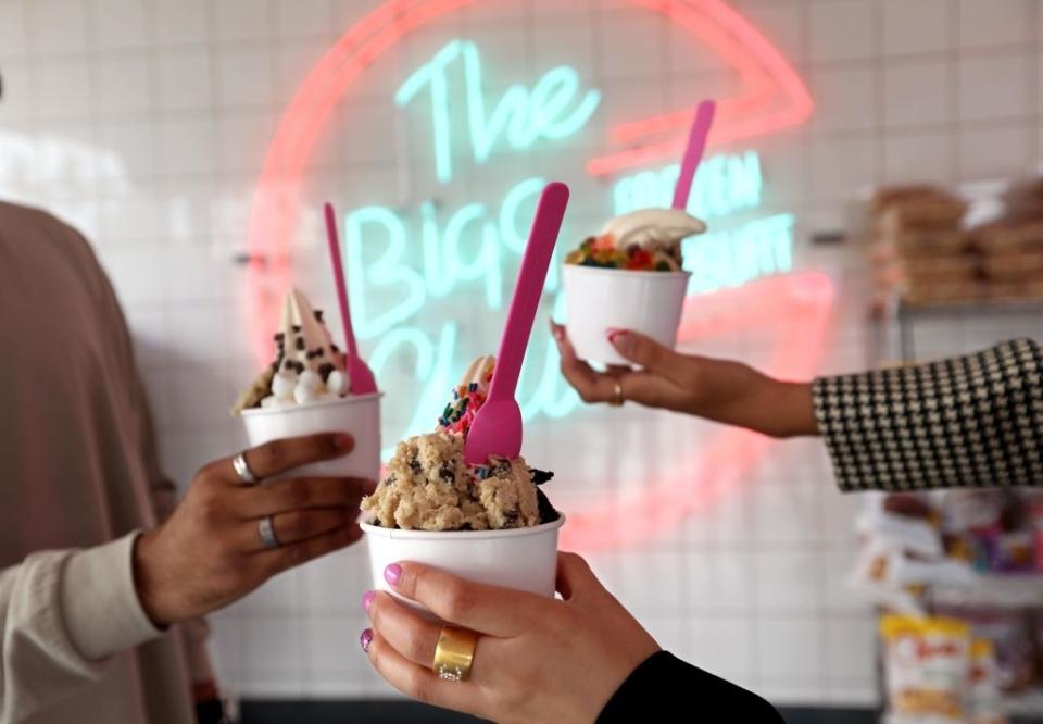 Two people holding cups of ice cream with toppings, neon sign in background reads "The Big O"