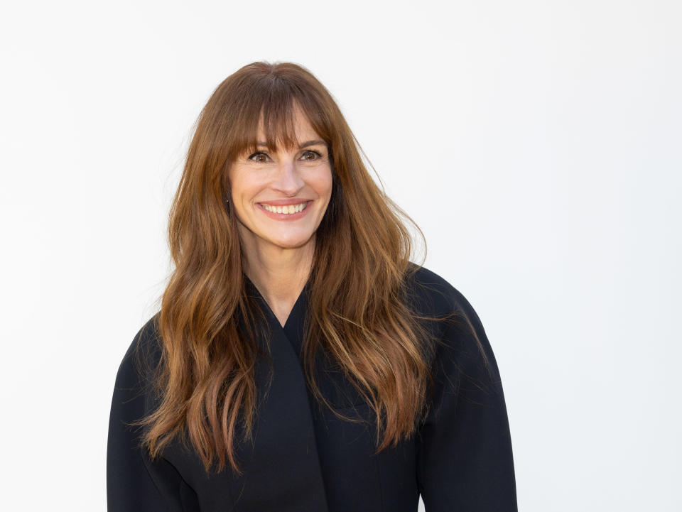 Julia Roberts smiling, wearing a dark outfit with her hair down, against a plain background