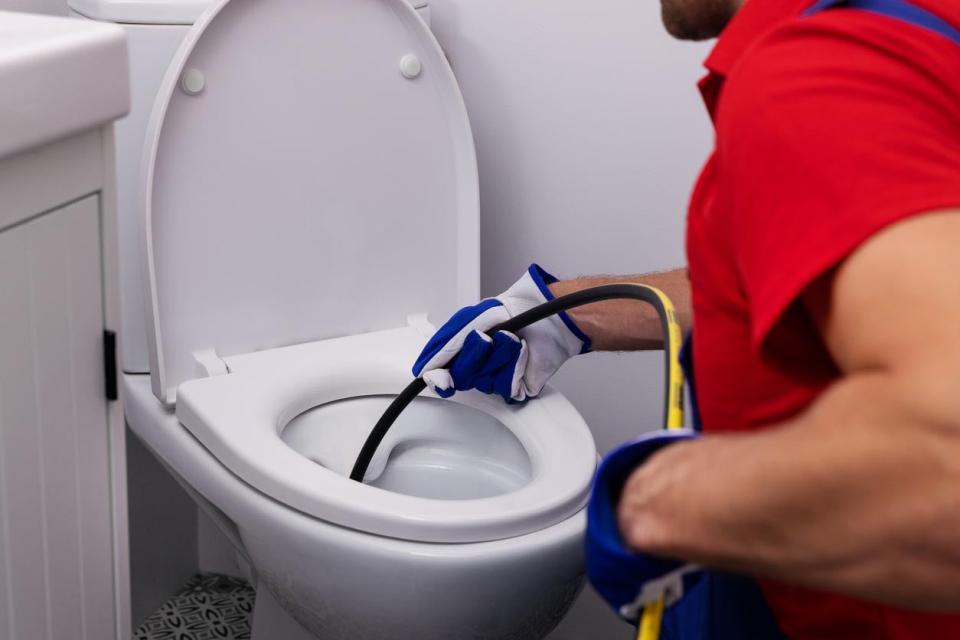 A man in a red shirt uses a tool to fix the inside of a toilet.