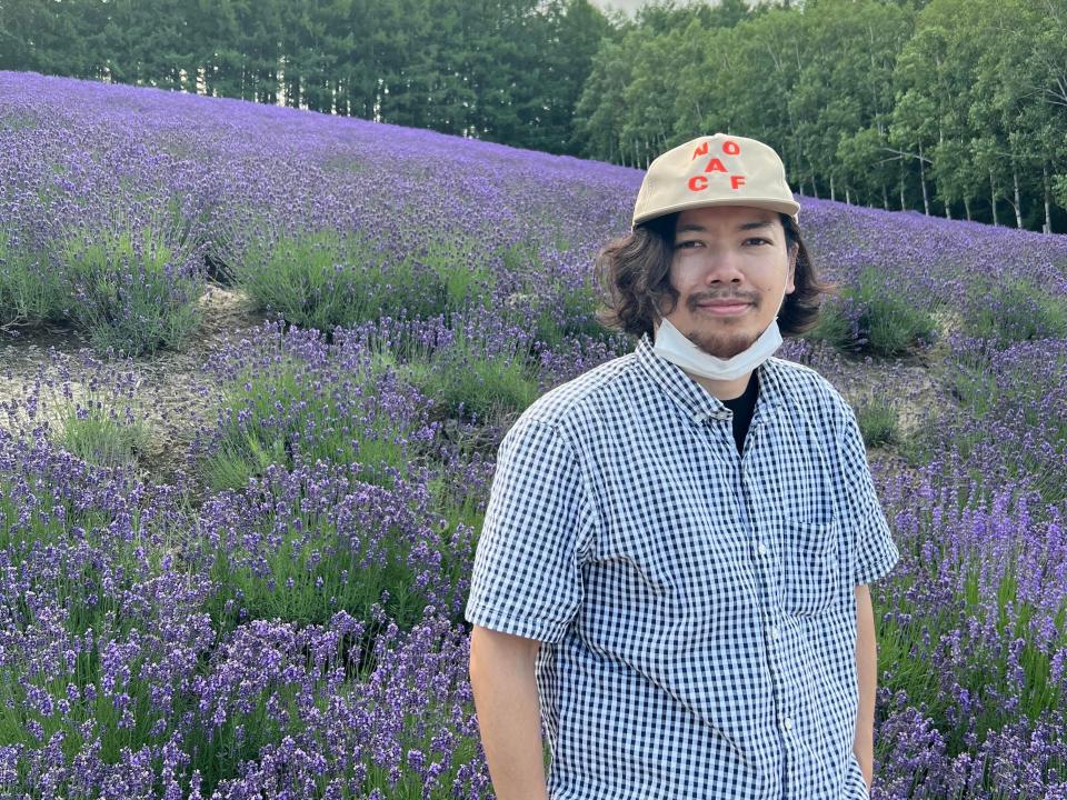 Isechi Makoto standing in front of a field of lavender while wearing a baseball cap and a white checkered shirt.