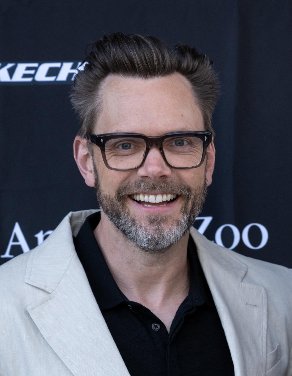 A closeup of Joel McHale smiling at a media event. He's wearing a casual suit and eyeglasses