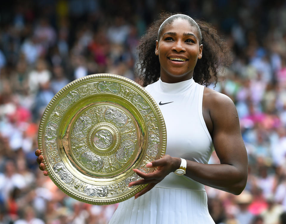 Serena Williams stands holding a large silver trophy at a tennis event, wearing a sleeveless white dress