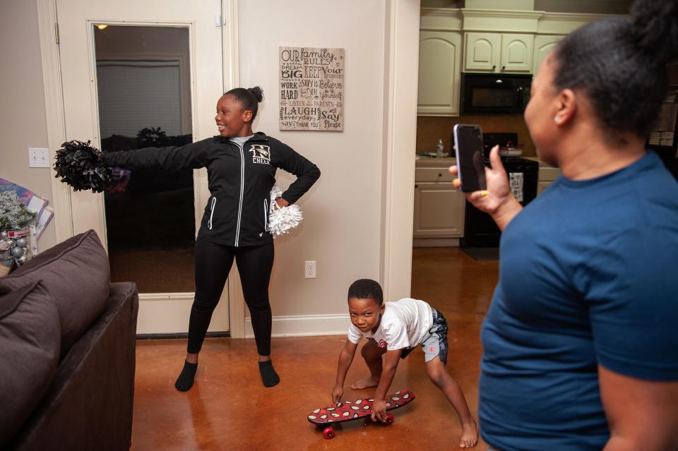 A woman holds up her phone to capture a girl practices a cheer routine and her boy playing with his skateboard.