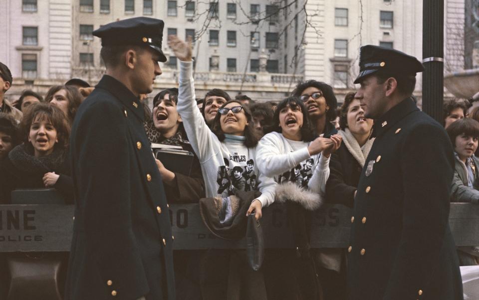 View of teenagers, some wearing band sweatshirts, and young female fans of English pop group The Beatles, shouting from behind a police line, along with NYPD police officers outside the Plaza Hotel