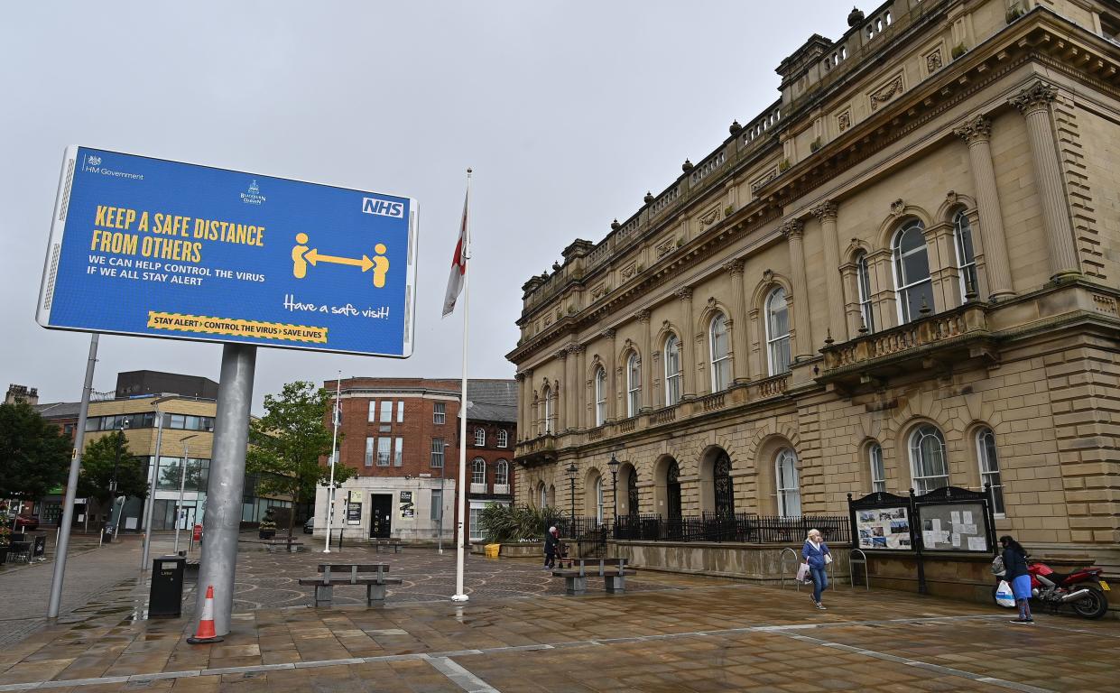 Pedestrians, some wearing a face mask or covering due to the COVID-19 pandemic, walk past an electronic sign alerting people to social distance due to COVID-19, in Blackburn, north west England, on July 15, 2020, following news that there has been a spike in the number novel coronavirus cases in the area. (Photo by Paul ELLIS / AFP) (Photo by PAUL ELLIS/AFP via Getty Images)