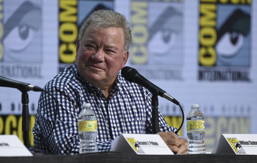 Man, William Shatner, in plaid shirt, sitting in front of mic on panel at comic con, looking to the left, smiling