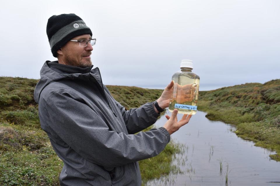 A scientist in a raincoat and cap holds up a water sample in a jar.