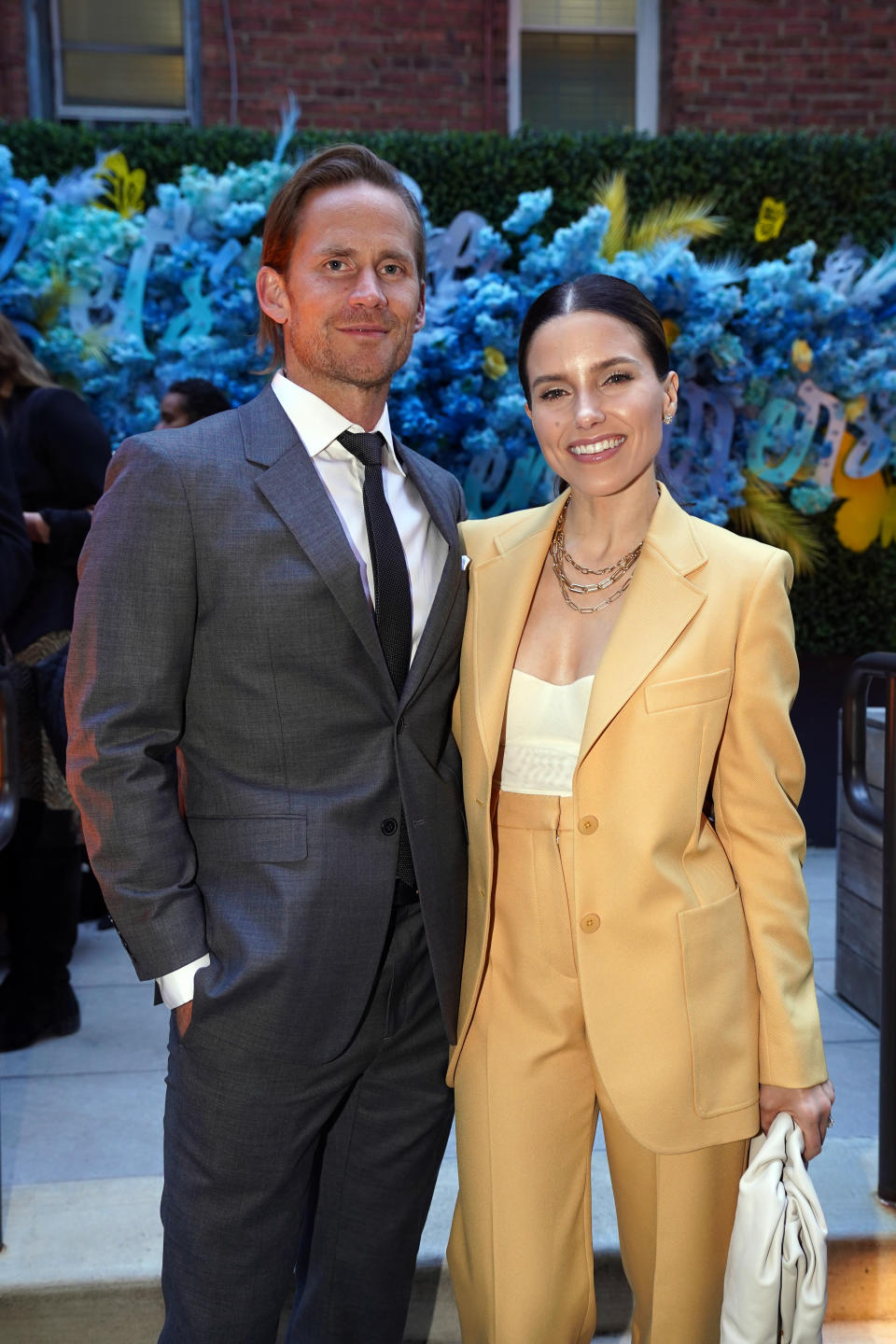 Grant Hughes and Sophia Bush smile for a photo at an event. Grant is wearing a suit and tie and Sophia is wearing a pantsuit