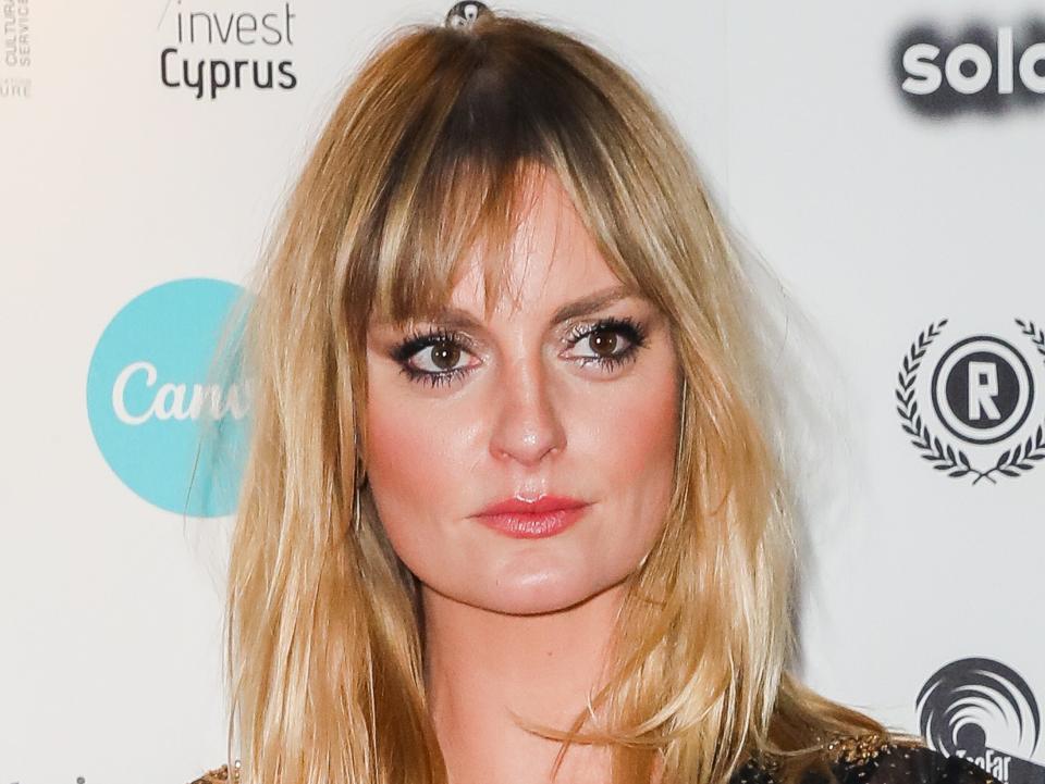 morgana robinson wearing a black dress and black and gold cardigan, standing on a red carpet and looking slightly to the left of the camera