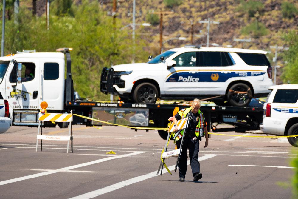 A police officer carries traffic barricades across 27th Avenue near Dear Valley Road, Sunday, Aug. 29, 2022, in Phoenix, as a damaged police vehicle is transported out of the area after a shooting occurred the night before injuring two Phoenix police officers. (Megan Mendoza/The Arizona Republic via AP)