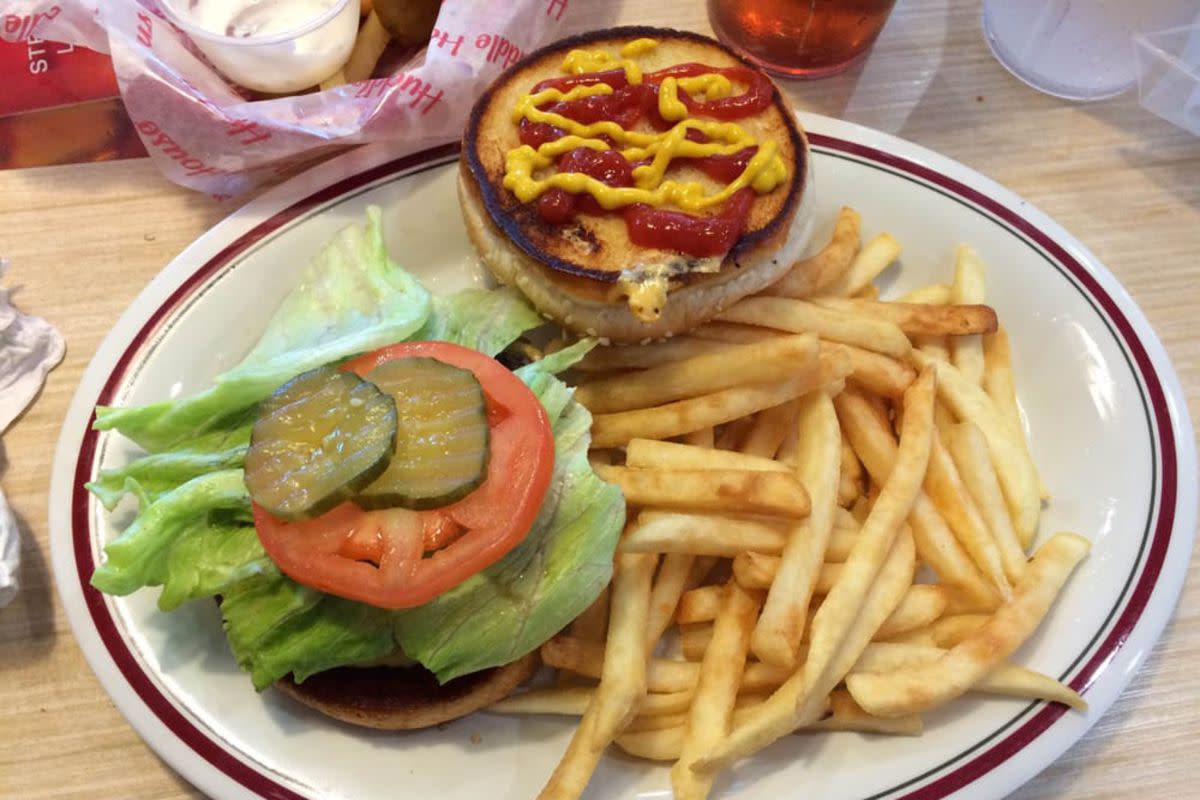 Huddle House hamburger and fries on a maroon-lined white plate, a drink and dipping sauces in the top background on a wooden table at Huddle House