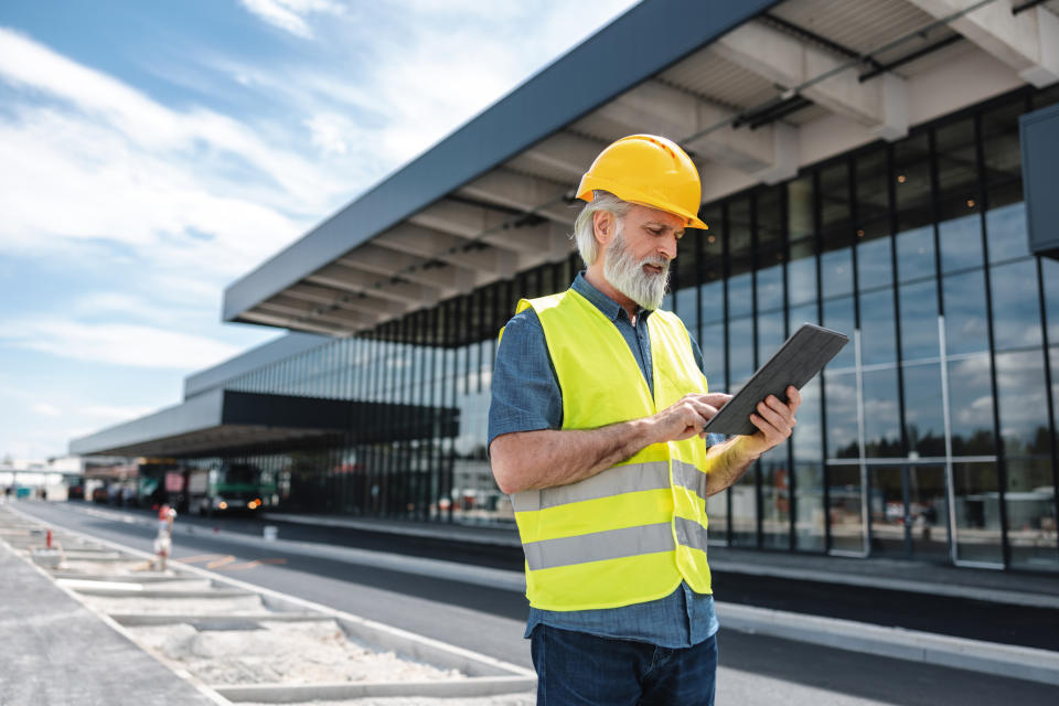 An older man wearing a yellow hard hat and reflective vest works on a tablet outside a modern commercial building under construction