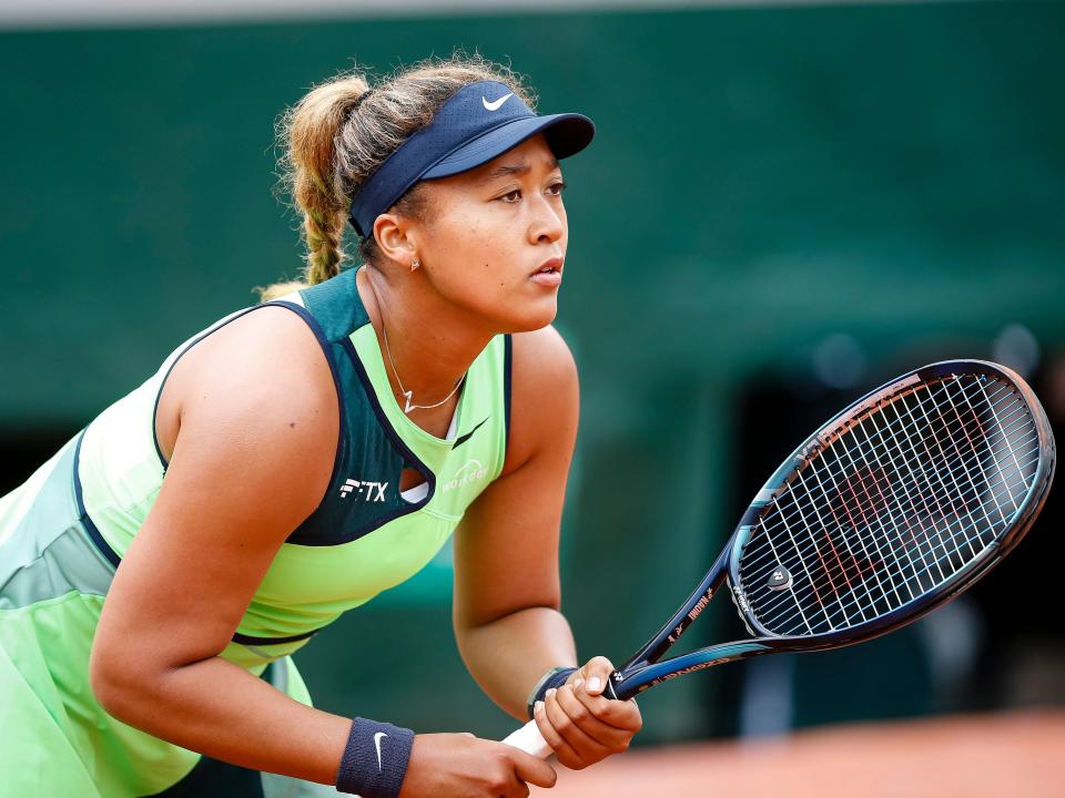 Naomi Osaka wears a green tennis outfit while holding a tennis racket and looking focused during a match.