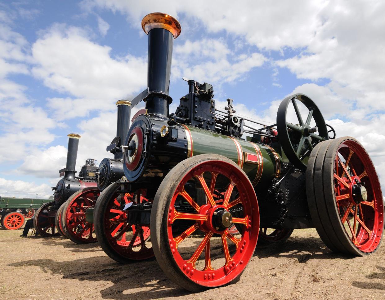 Several large red and green traction engines in a row with a blue, cloudy sky