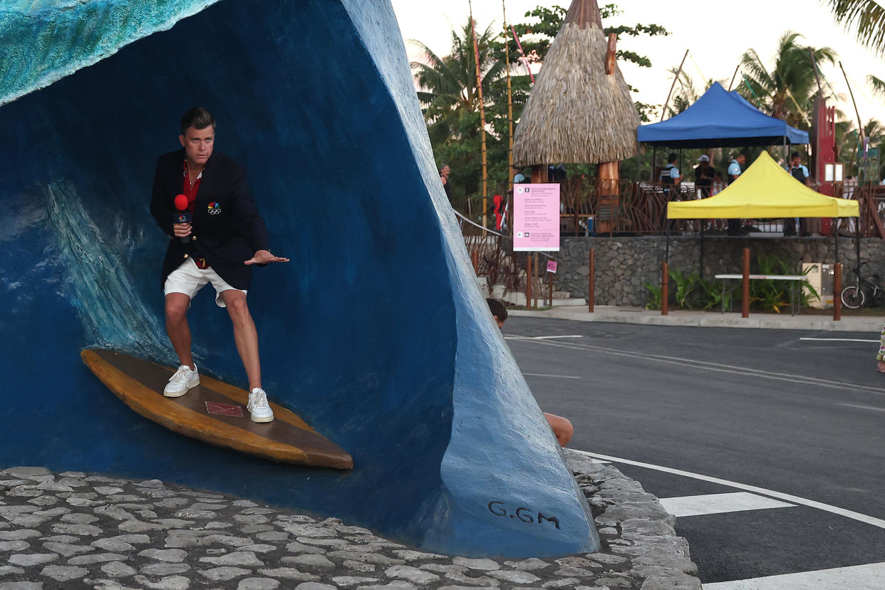 Colin Jost holds a microphone and stands on a prop fashioned to look like a surfboard on a wave as he films a segment as he covers Olympic surfing in Tahiti for NBC.