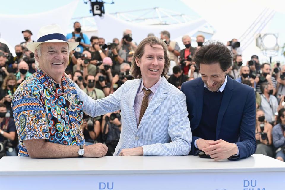 bill murray, wearing a patterned button shirt and white fedora, smiles as wes anderson, wearing a light blue suit, places a hand on his shoulder, with multiple photographers taking pictures behind them