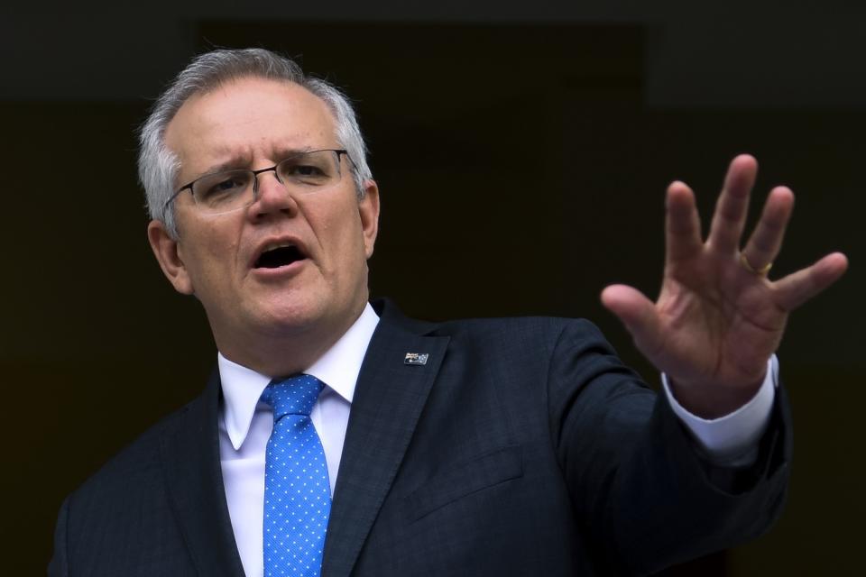 A man in dark suit and blue tie gestures with one hand as he speaks.