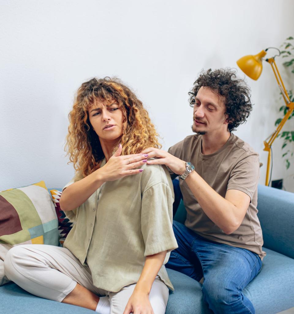 A woman and a man sit on a couch; the man appears to massage the woman's shoulder as she looks at her back with discomfort