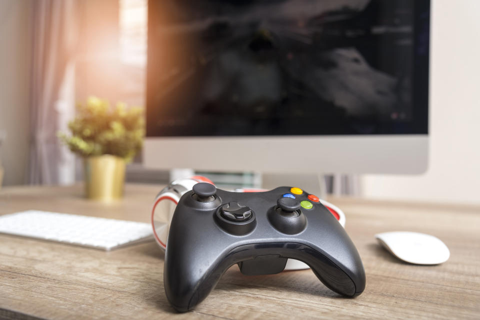 A black video game controller sitting on a table with a mouse, keyboard, and PC monitor in the background.