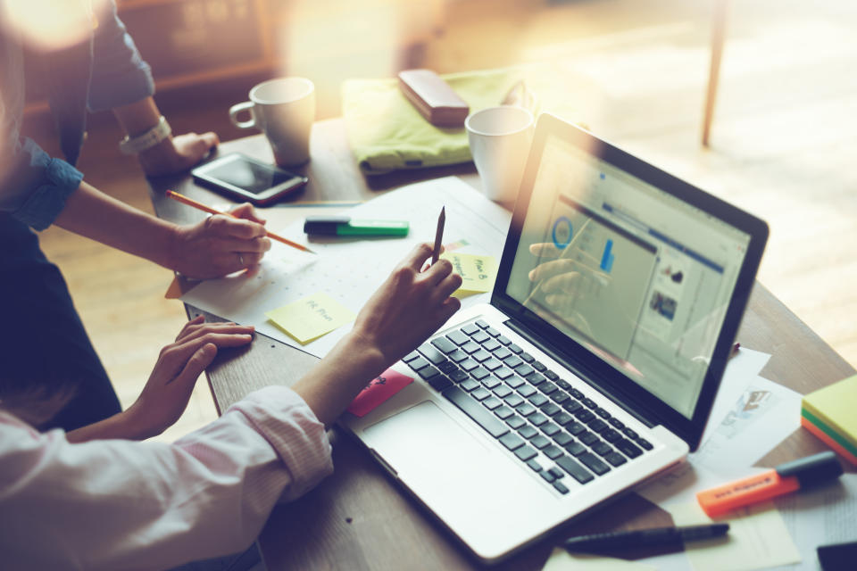 Two people working on market research on a notebook computer with papers covering desk.