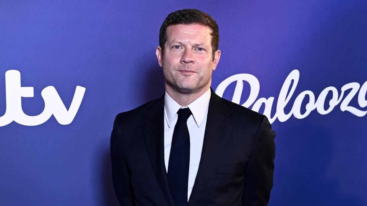  Dermot O'Leary standing in front of a purple background wearing a dark suit and tie. 