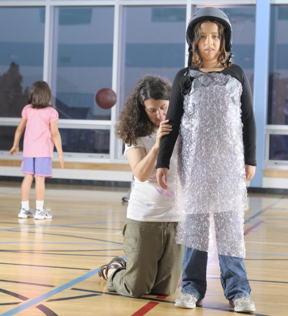 mom bubble wrapping her kid, who wears a helmet, before they can play in a gymnasium