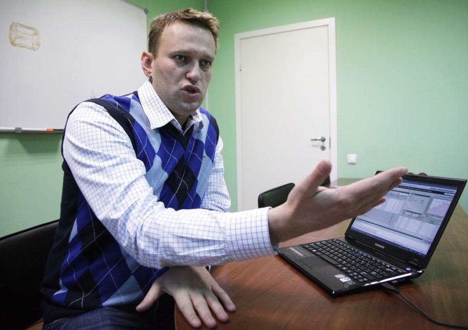 alexei navalny speaks while sitting at a table inside a room with green walls, a computer is open on the table in front of him and he holds one hand out, he wears a blue sweater vest over a patterned collared shirt, a door and hanging dry erase board are behind