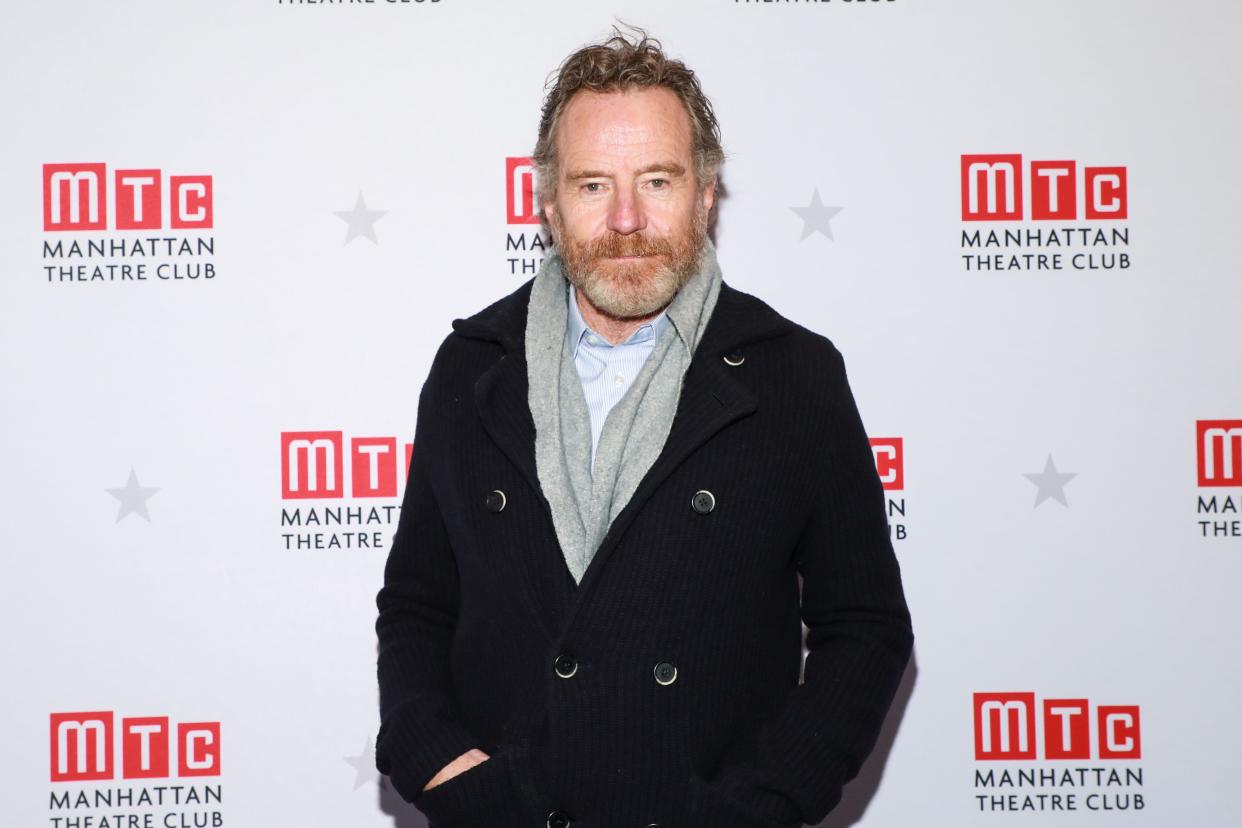 Bryan Cranston wears a dark peacoat  and a grey scarf in front of a white backdrop that reads "Manhattan Theatre Club" in New York City.