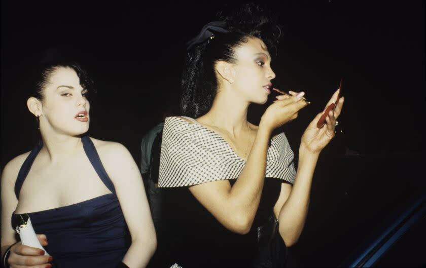 Carmen Xtravaganza stands next to another person, wearing a black outfit, staring into a compact mirror, applying makeup
