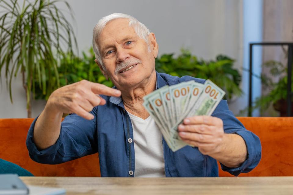 Smiling person sitting at a table and pointing to a stack of $100 bills in their hand.