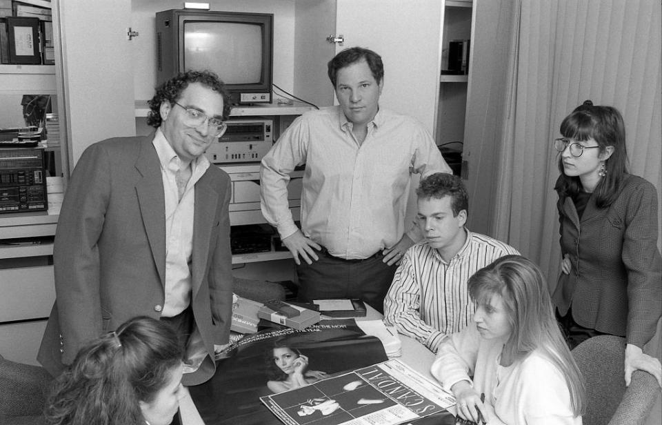 bob weinstein and harvey weinstein stand behind a table and look at the camera, three people are seated at the table and another woman stands to the right of the table, a tv and other equipment is in a cabinet behind them and movie posters are on the table