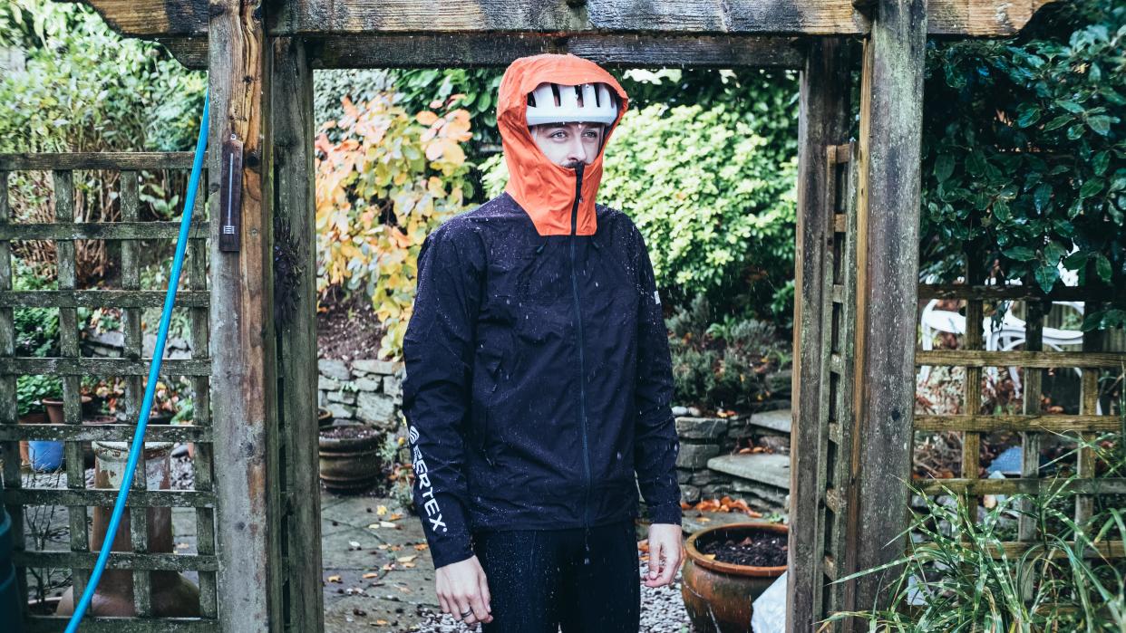  A white man wears a waterproof jacket under a stream of water in a garden 