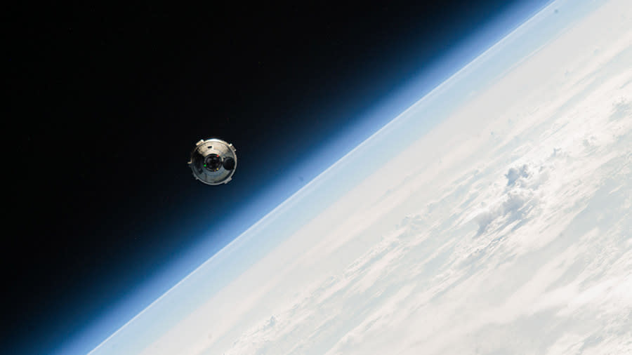  A silvery white space capsule hovers in orbit with earth in the background. 