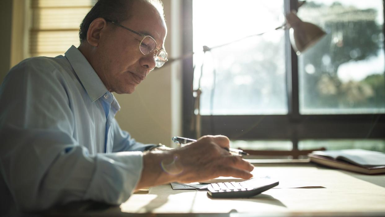 Senior Taiwanese man with eyeglasses sitting in dining room at home and calculating bills for paying.