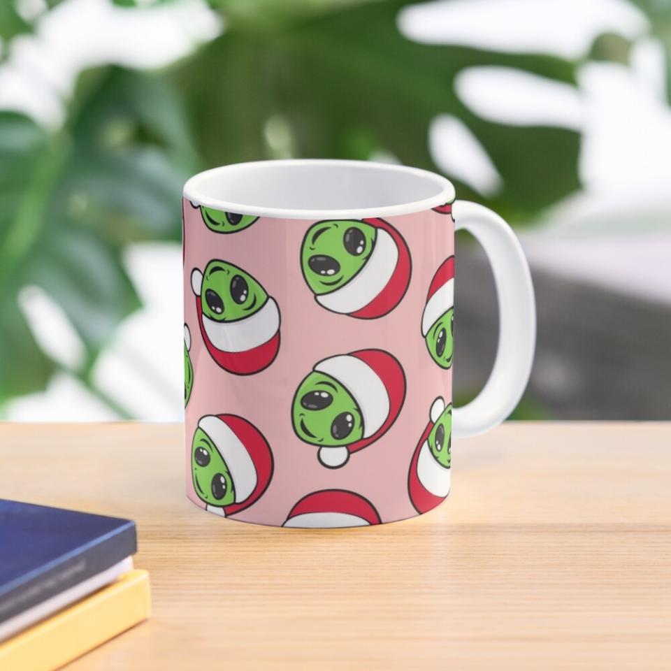 A mug with a pink background and green Christmas aliens wearing Santa hats on it