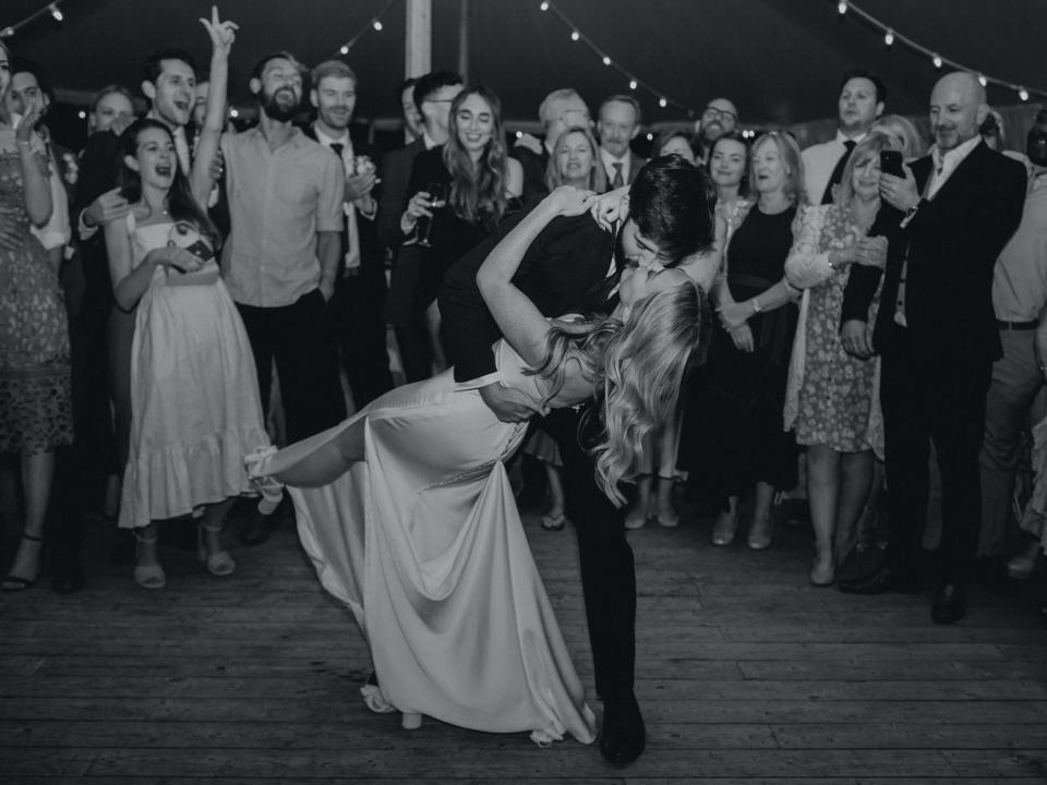 A groom dips and kisses his bride as their wedding guests look on and clap.