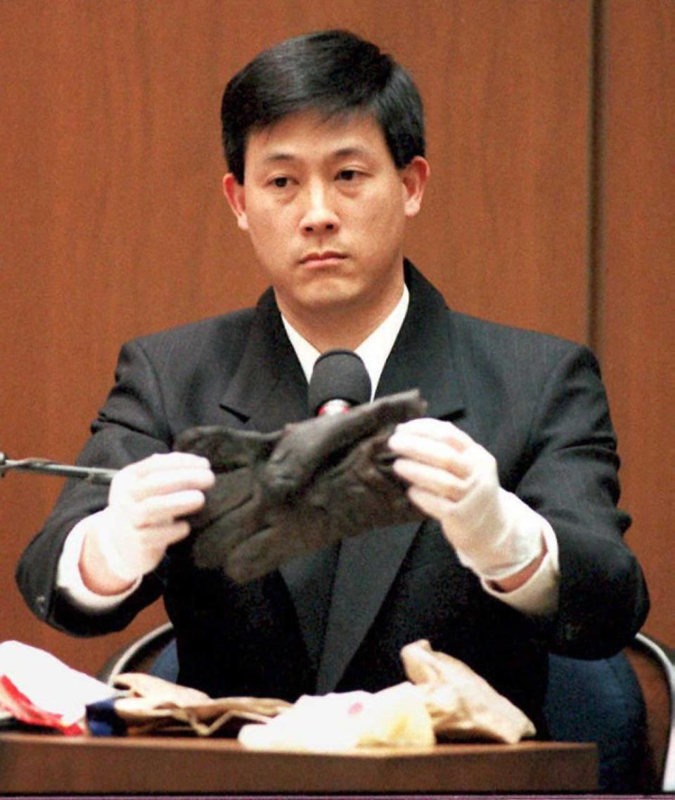 dennis fung sits on the witness stand and holds a black glove with his hands in white gloves, he wears a dark suit jacket and white collared shirt