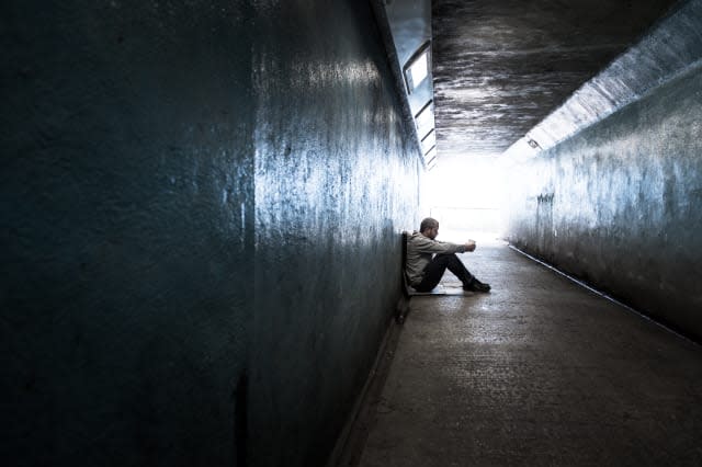 Wide angle image of a young homeless adult male sitting on the hard floor of a subway tunnel. He is holding a paper cup and begg