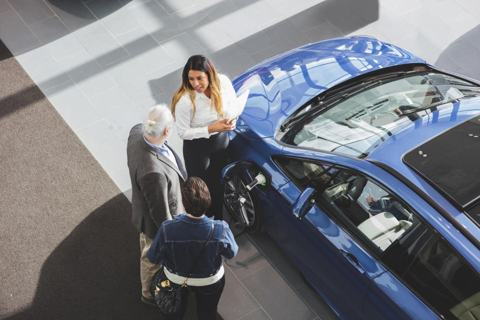 Three people, including a car salesperson, discuss near an electric car in a showroom