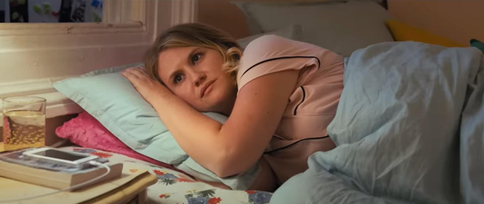 Jillian Bell as Brittany Forgler is lying in bed, resting her head on her hand and looking contemplative. A glass of water, a book, and a phone are on the bedside table nearby.