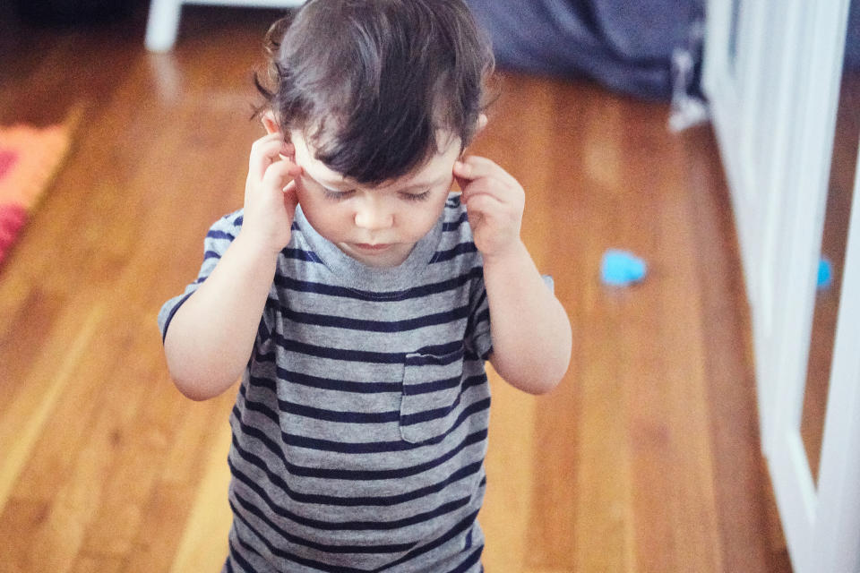 A young child stands indoors, wearing a striped shirt, with their hands up to their ears, appearing to be either listening intently or annoyed by a sound