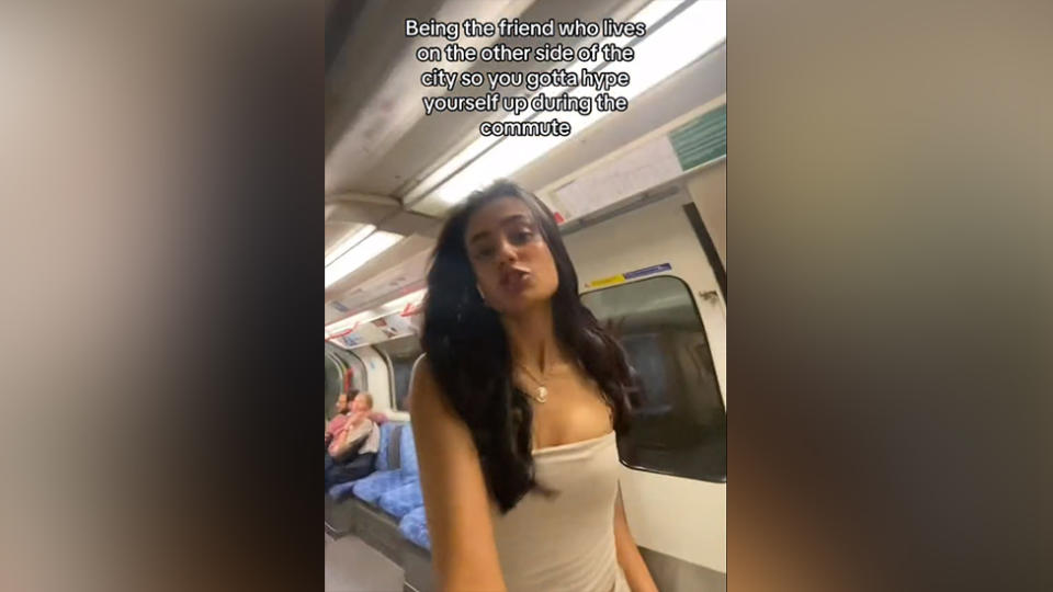 A still from a self-shot video of a young Asian woman dancing in a Tube carriage. She's about to flip her long black hair to the side. She's wearing a white tube top, and her long black hair cascades down her shoulders. Behind her, other passengers are sitting down as normal. A caption is embedded on the video which reads: "Being the friend who lives on the other side of the city so you gotta hype yourself up during the commute".
